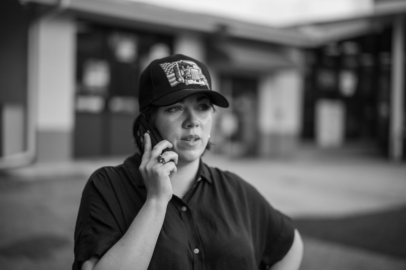 A woman wearing a dark shirt and an American flag-patterned baseball cap talks on a mobile phone in front of a blurry industrial building in this black-and-white medium shot.