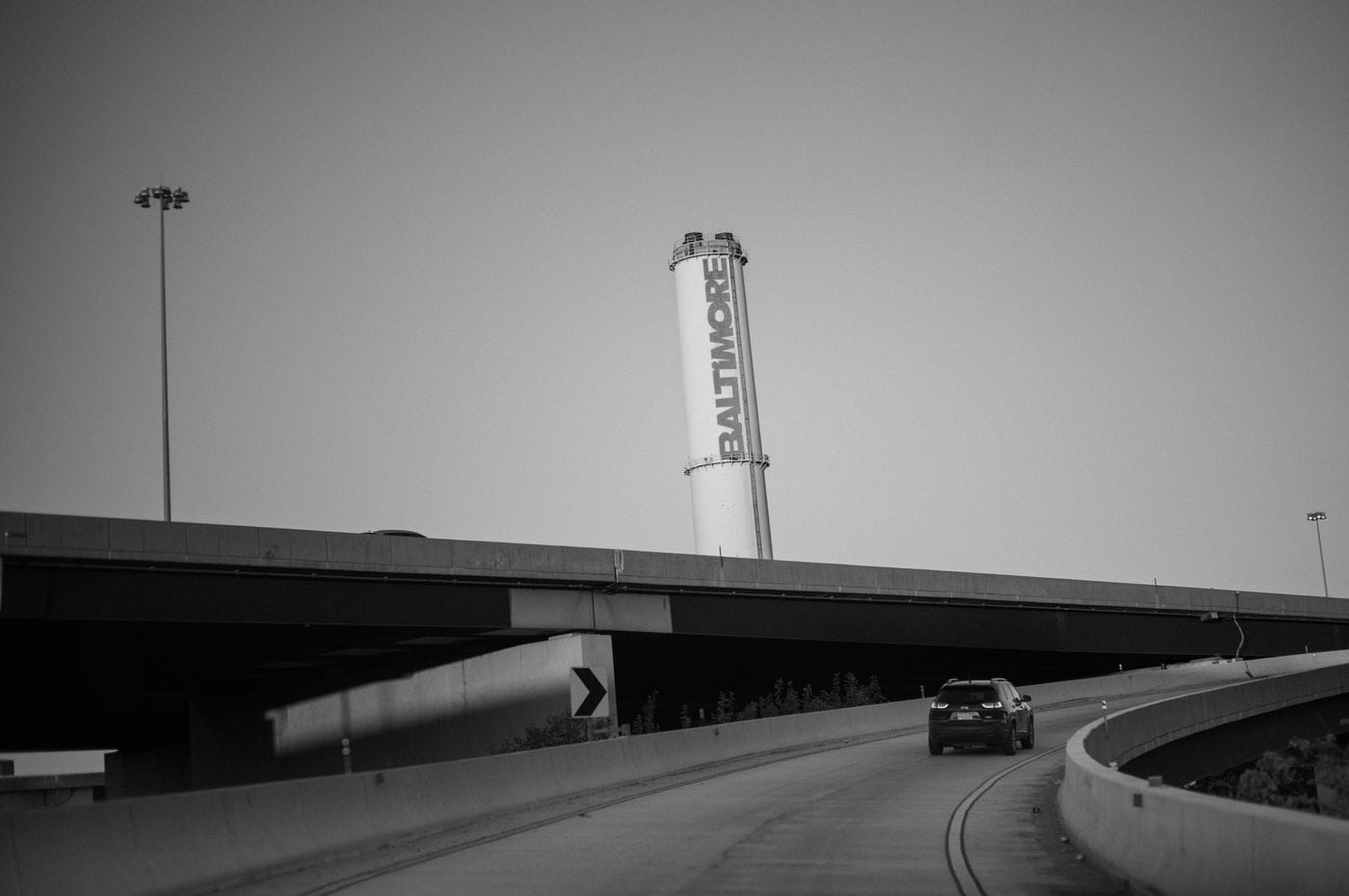 A black-and-white view of a car driving on a highway ramp toward a tall, cylindrical industrial tower emblazoned with the word "BALTIMORE."