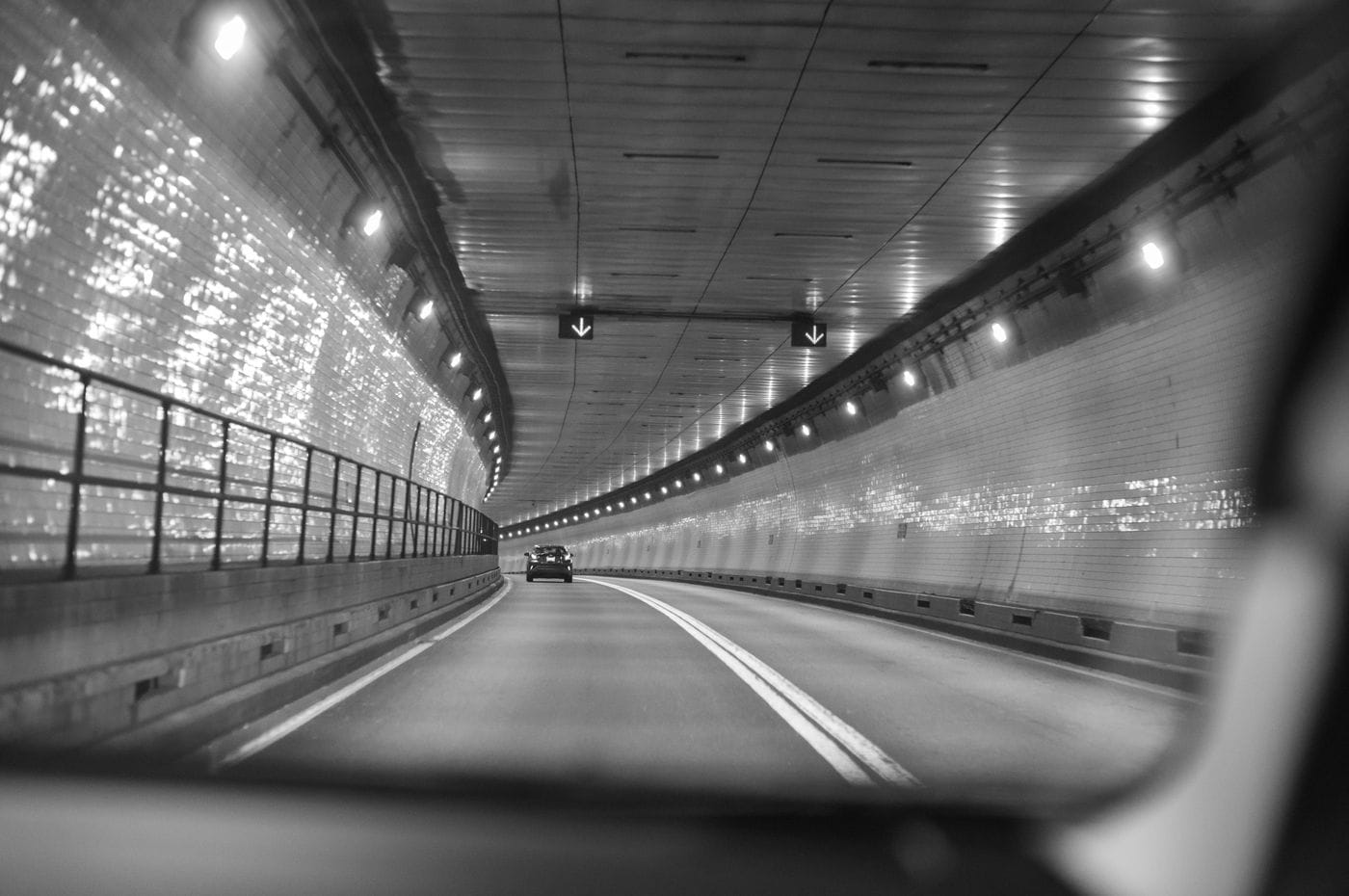 A view from inside a vehicle driving through a curved tunnel with tiled walls, bright overhead lights, and a lone car traveling in the distance.