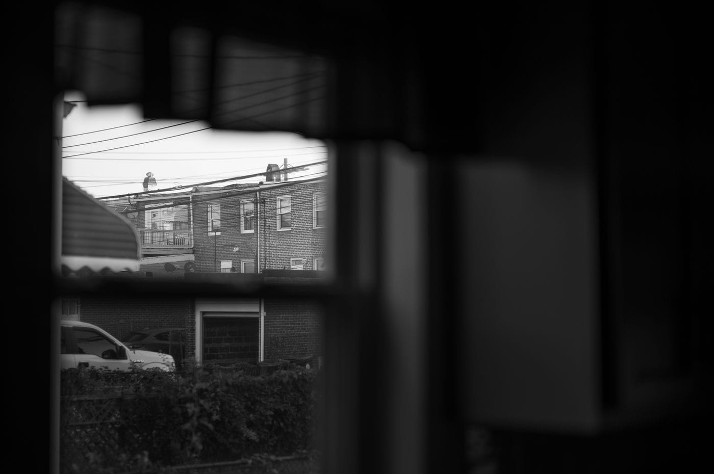 A black-and-white view through a window reveals a brick building with several windows, a balcony, and power lines against a bright sky, overlooking a parked car and overgrown foliage.