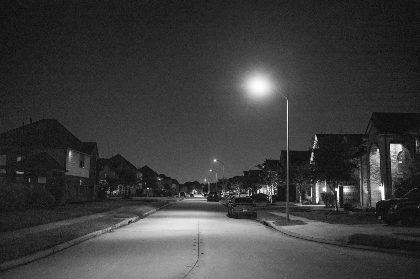 A black and white photo of a quiet residential street at night, illuminated by several glowing streetlights with houses and parked cars lining both sides.
