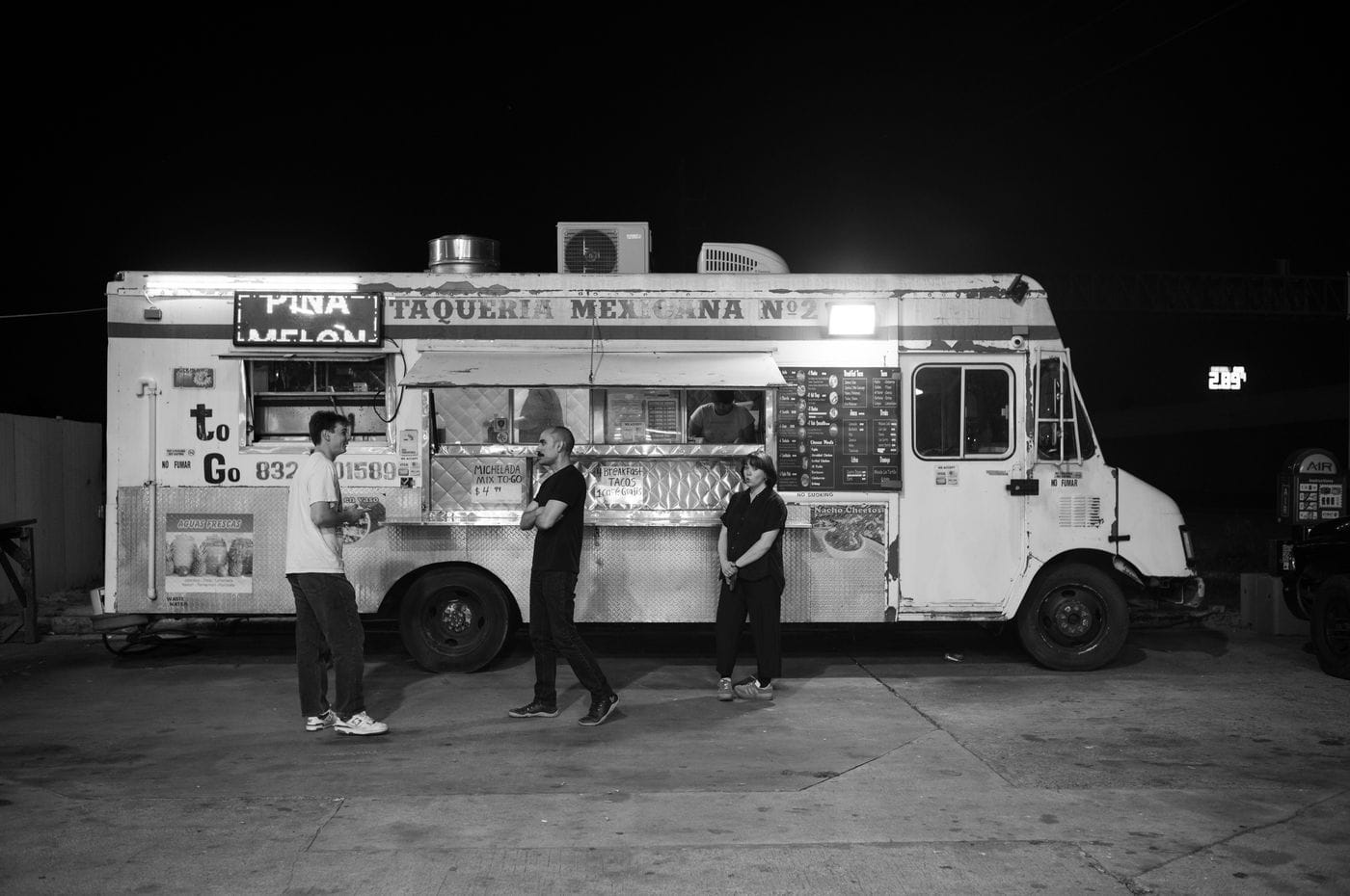 A black-and-white eye-level shot captures three people standing outside a parked, illuminated "Taqueria Mexicana No. 2" food truck at night, with one man standing to the left and another man and a woman standing closer to the service window.