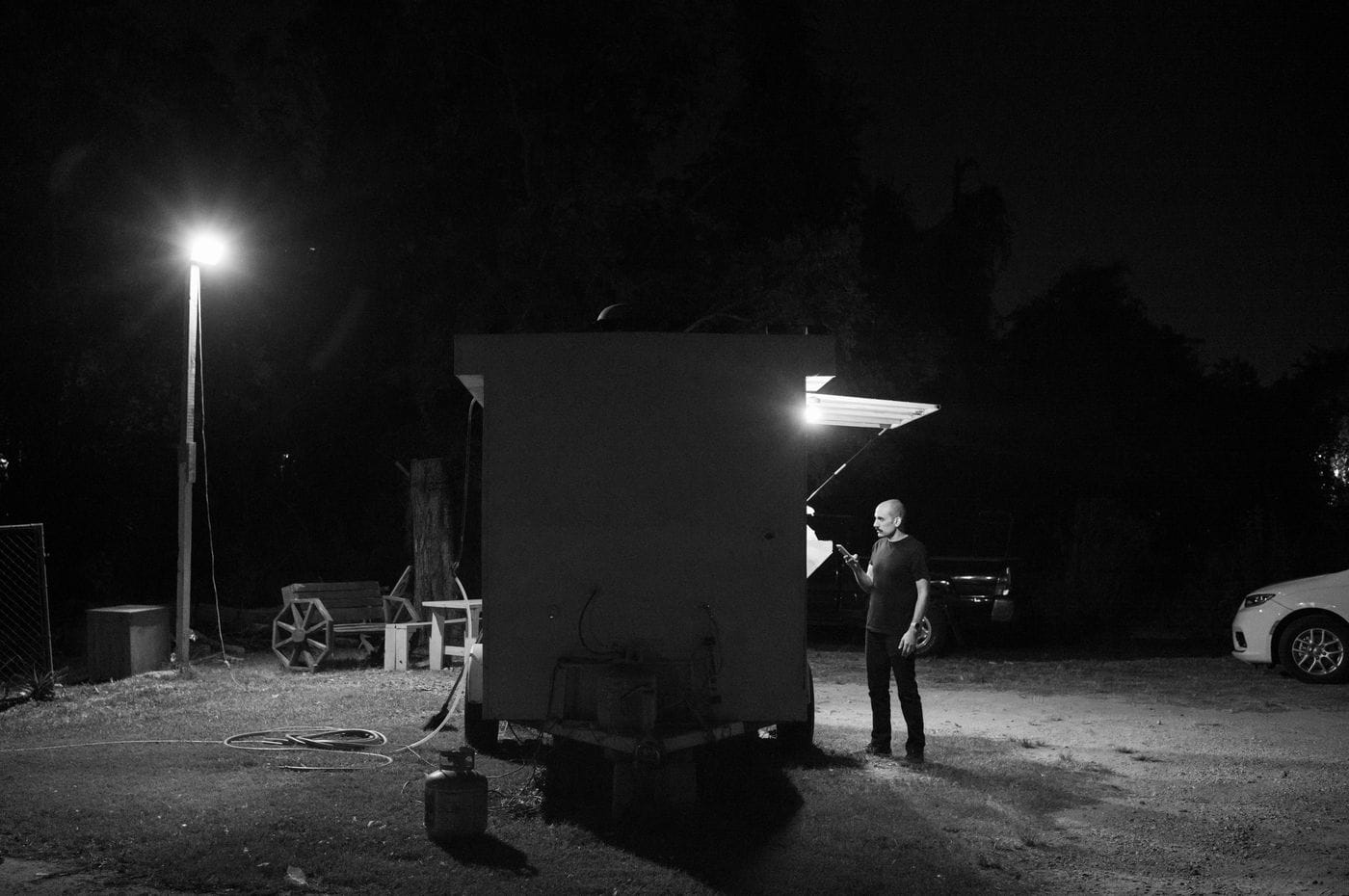 A bald man stands beside a small, illuminated food trailer at night, looking at his phone while a bright pole light shines on the grass nearby.