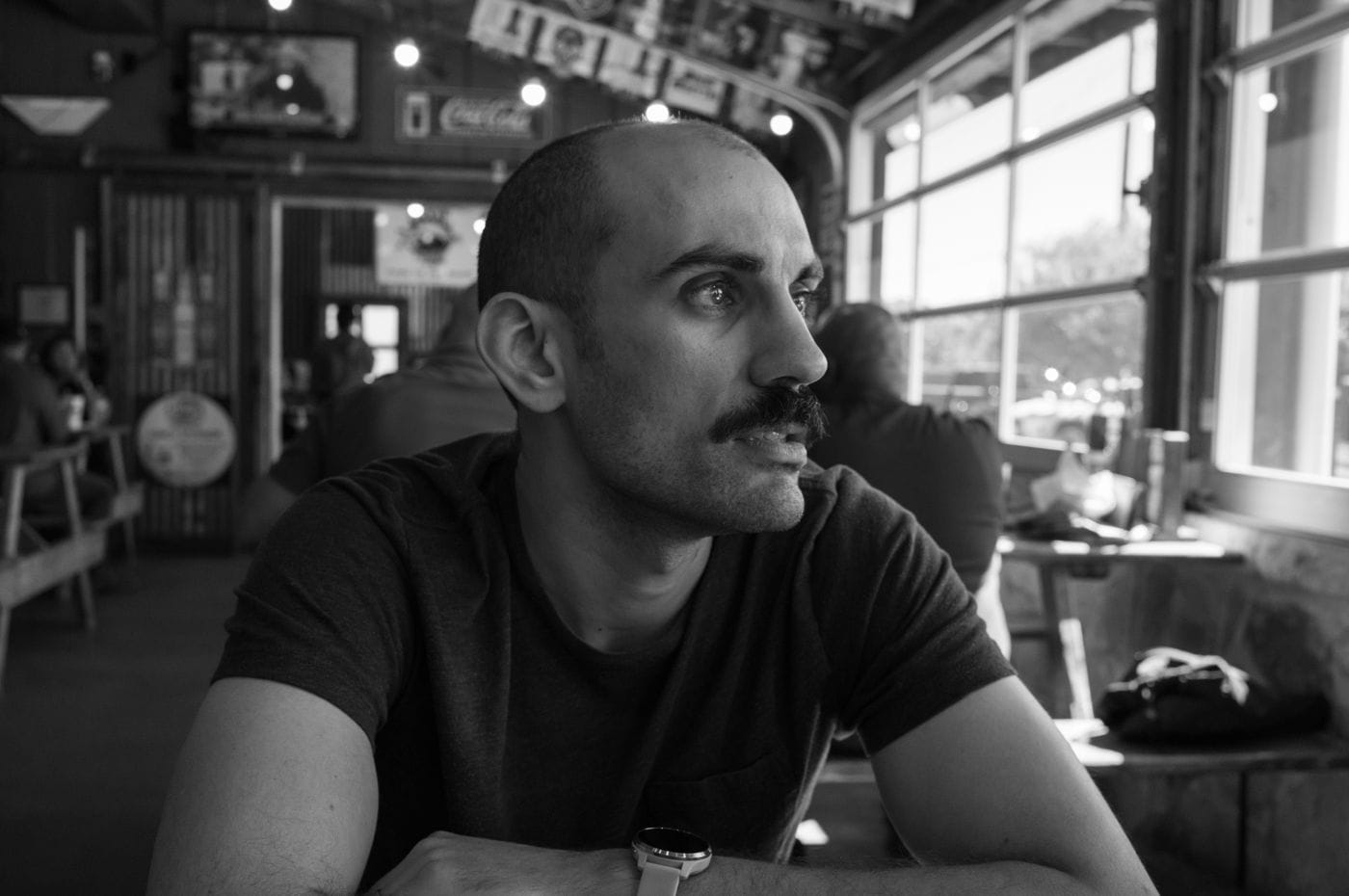 A medium-shot, black-and-white portrait of a man with a shaved head and a mustache, sitting at a table in a pub and looking off to the side.