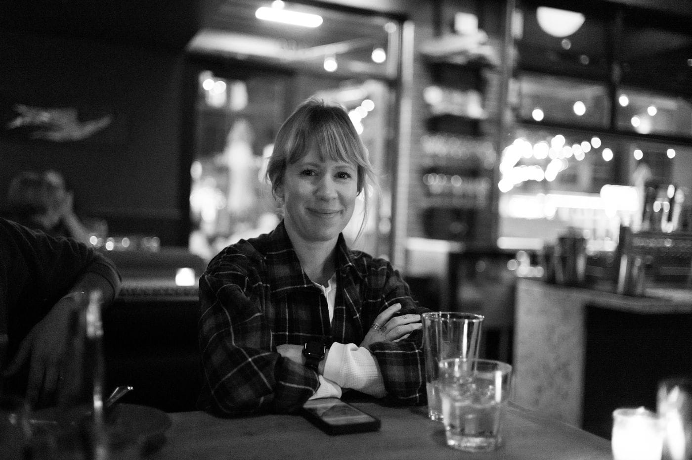 A woman with bangs and a plaid shirt sits at a table with her arms crossed in a dimly lit, out-of-focus restaurant setting.