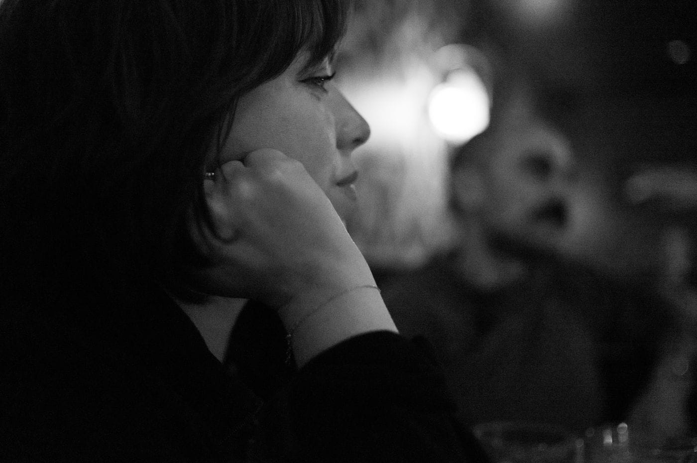 A side-profile, black-and-white close-up of a person with dark hair resting their chin on their hand, with a blurred silhouette of another person in the background.