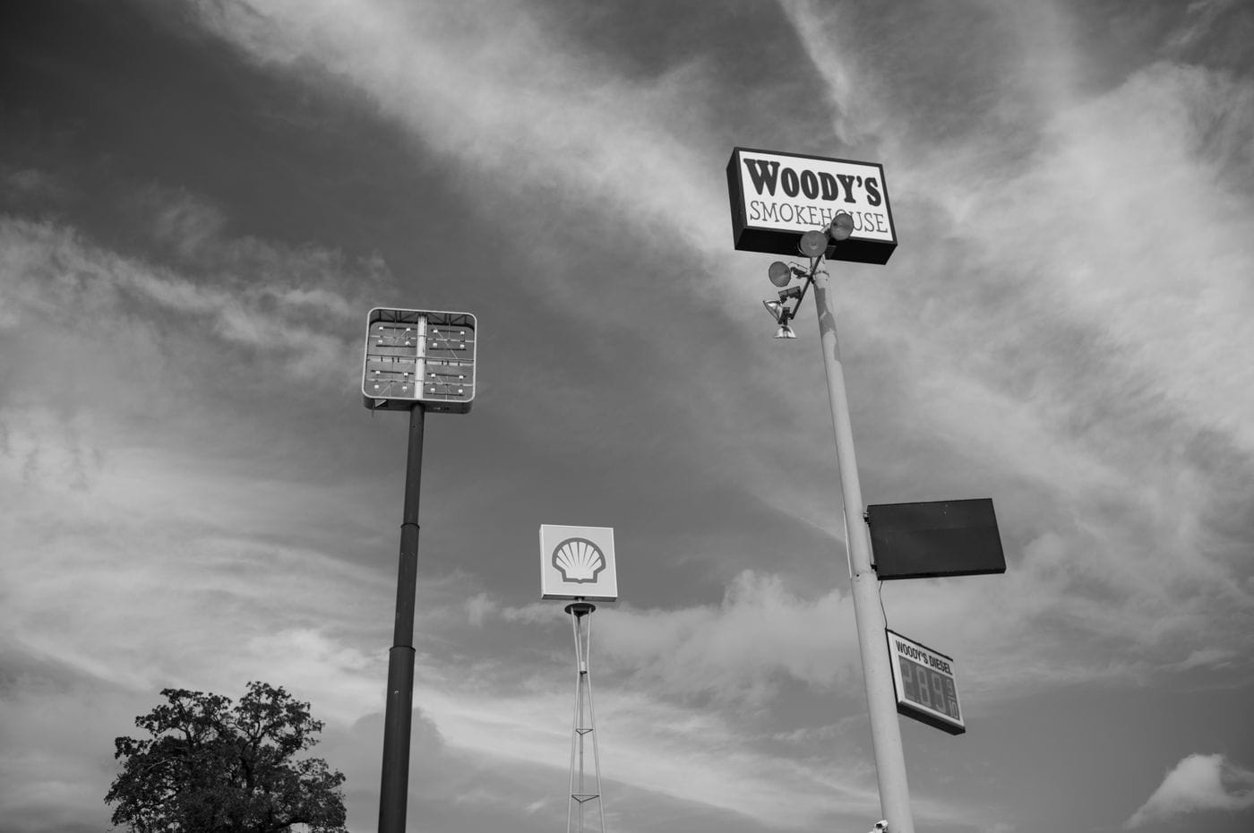 Low-angle black-and-white view of tall signage poles against a cloudy sky, including a Woody's Smokehouse sign, a Shell logo, and a stadium light fixture.
