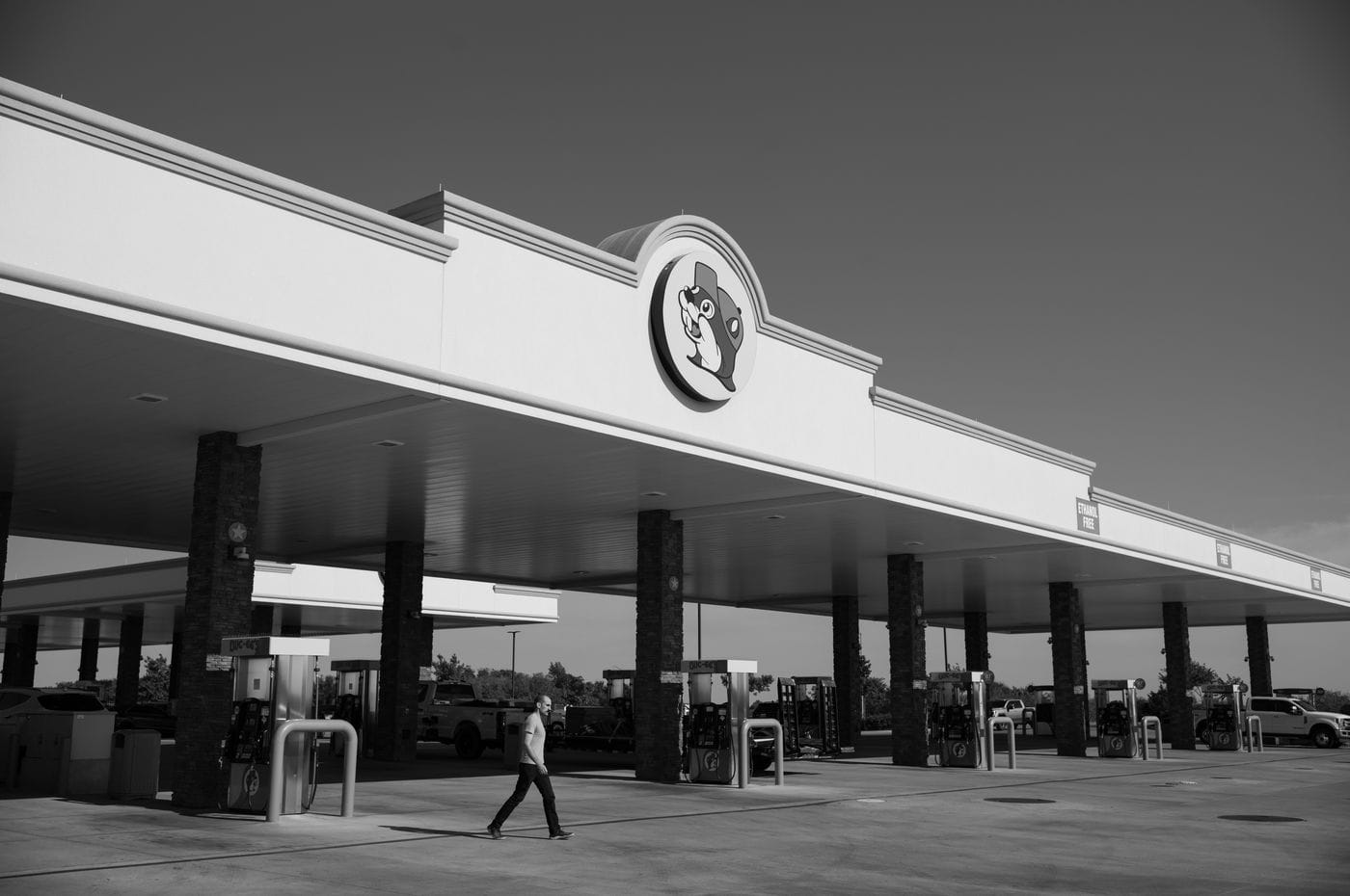 A man walks across the wide, concrete expanse of a large, sunny Buc-ee's gas station with stone-clad columns beneath a long, white, modern canopy.