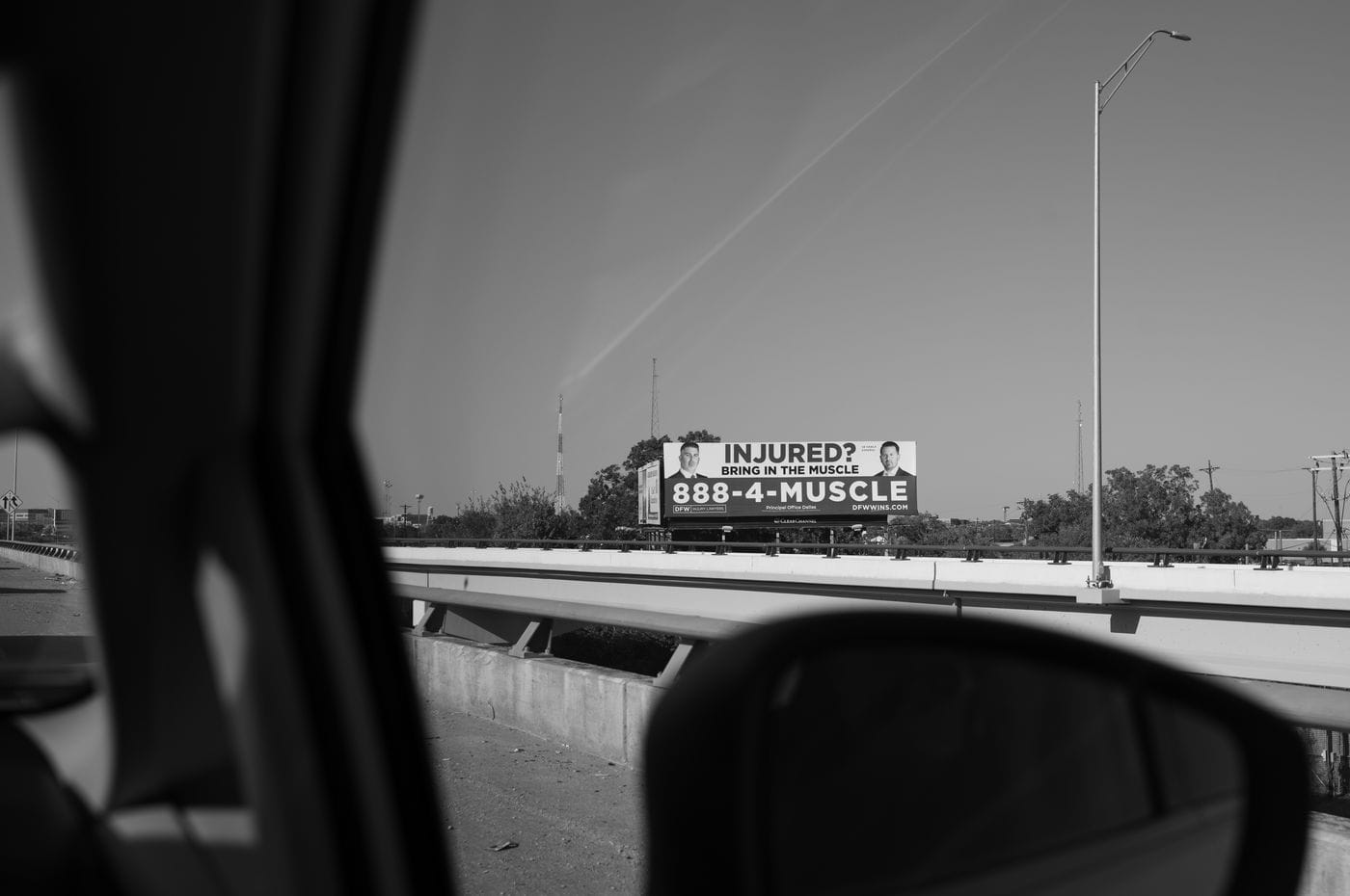 A billboard advertisement for injury lawyers featuring the phrase "INJURED? BRING IN THE MUSCLE" and the phone number "888-4-MUSCLE" is visible above a highway overpass, as seen from a vehicle window.