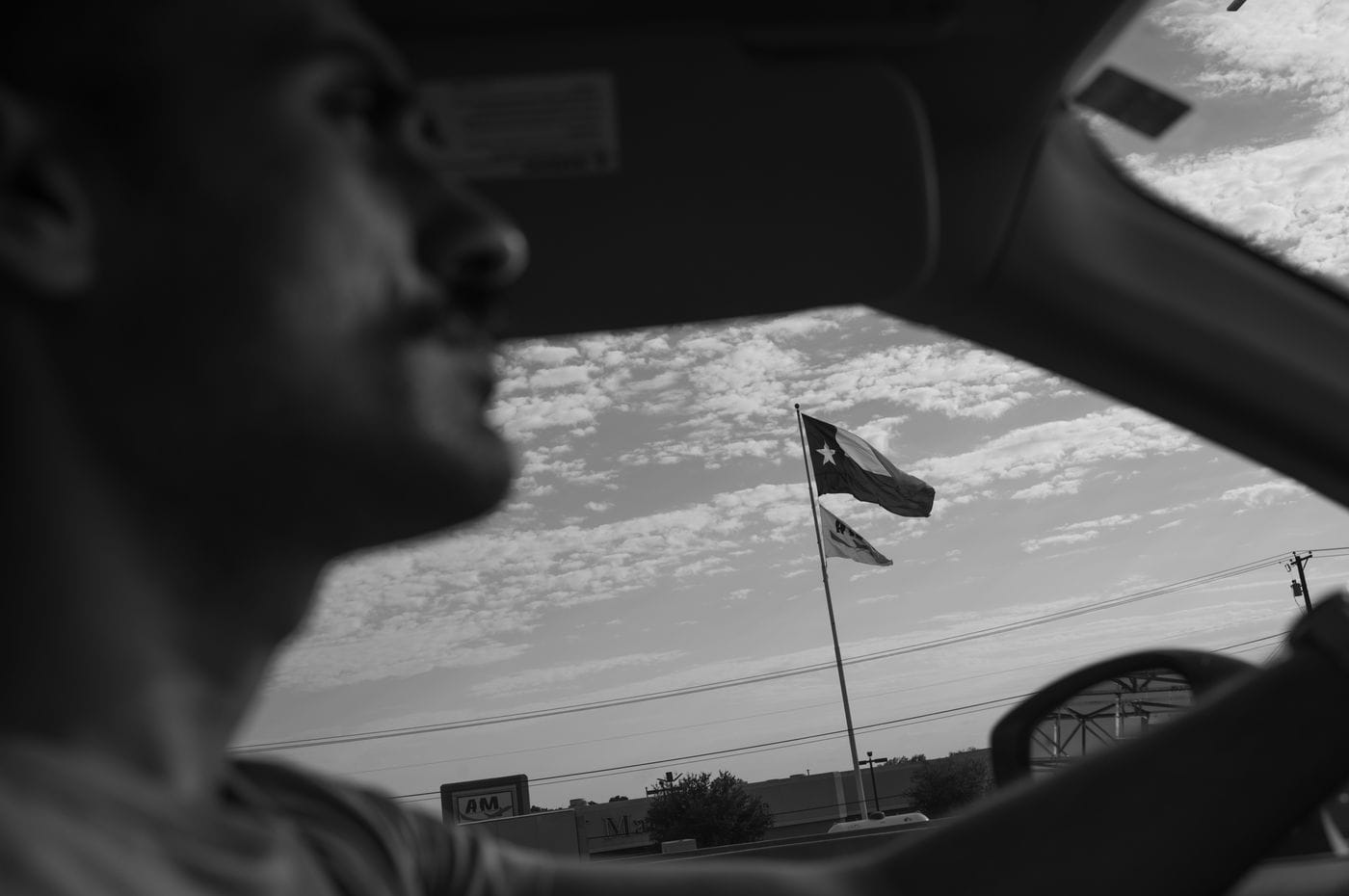 A black-and-white, through-the-windshield view shows a blurred driver in the foreground and a Texas state flag flying against a cloudy sky in the distance.