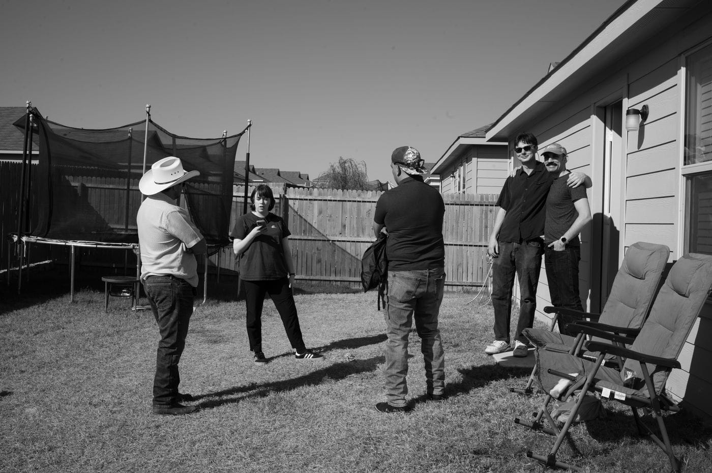 A group of five people stand in a sunny backyard, with a man in a cowboy hat on the left, a woman looking at her phone, a man with a backpack, and two men standing together in front of the house siding on the right.