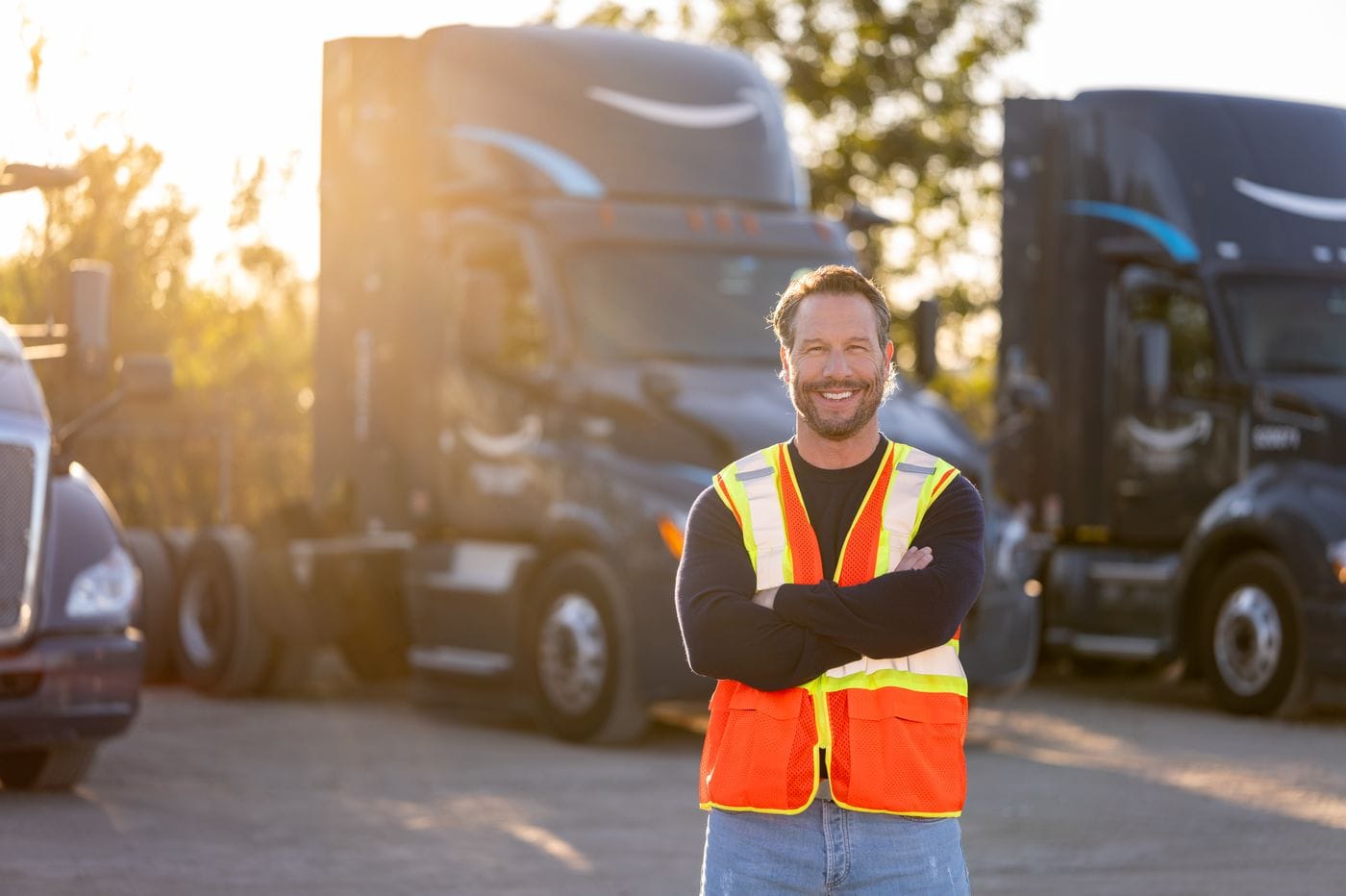 A middle-aged man with a beard, wearing a dark sweater, blue jeans, and a bright orange high-visibility safety vest, stands with his arms crossed in front of three parked semi-trucks at sunset.