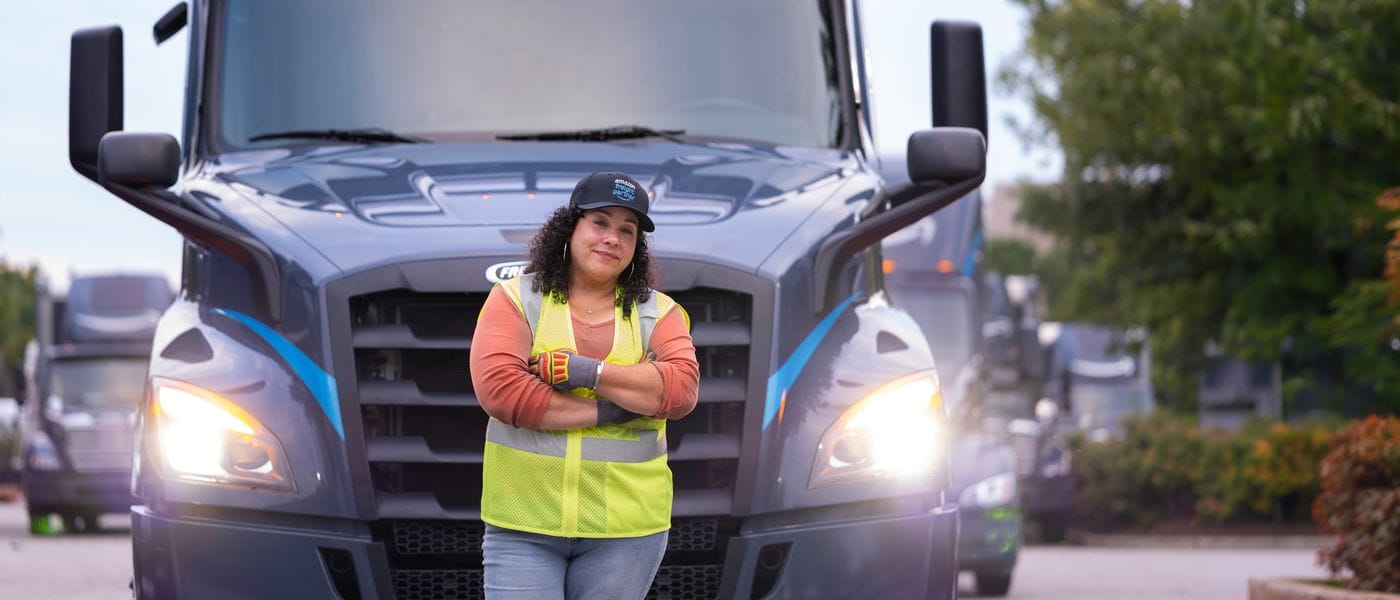 A female truck driver wearing a high-visibility yellow vest and an Amazon Freight Partner cap stands with her arms crossed in front of a parked dark gray semi-truck.