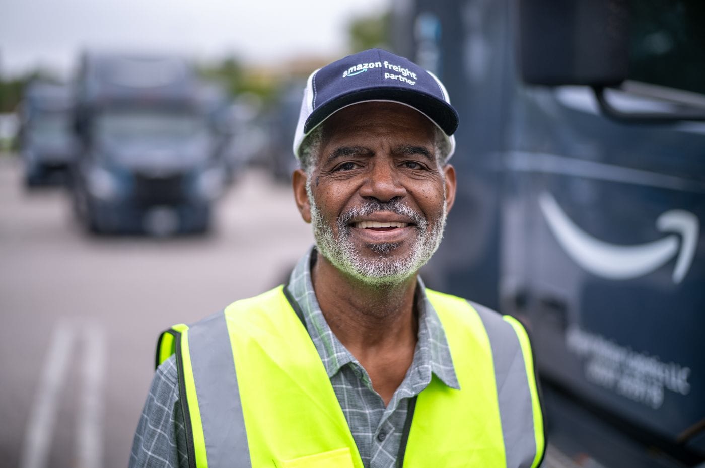 An older Black man with a graying beard and a bright yellow high-visibility vest stands outdoors wearing an Amazon Freight Partner cap, with a large blue freight truck visible in the background.