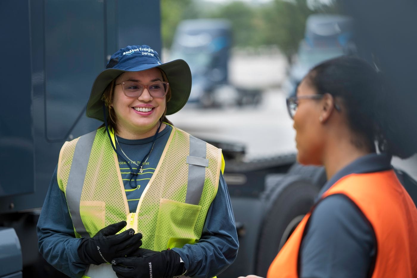 Two warehouse employees, one wearing a blue "Amazon Freight Partner" hat and a yellow safety vest, stand outdoors near a semi-truck while smiling and talking to each other.