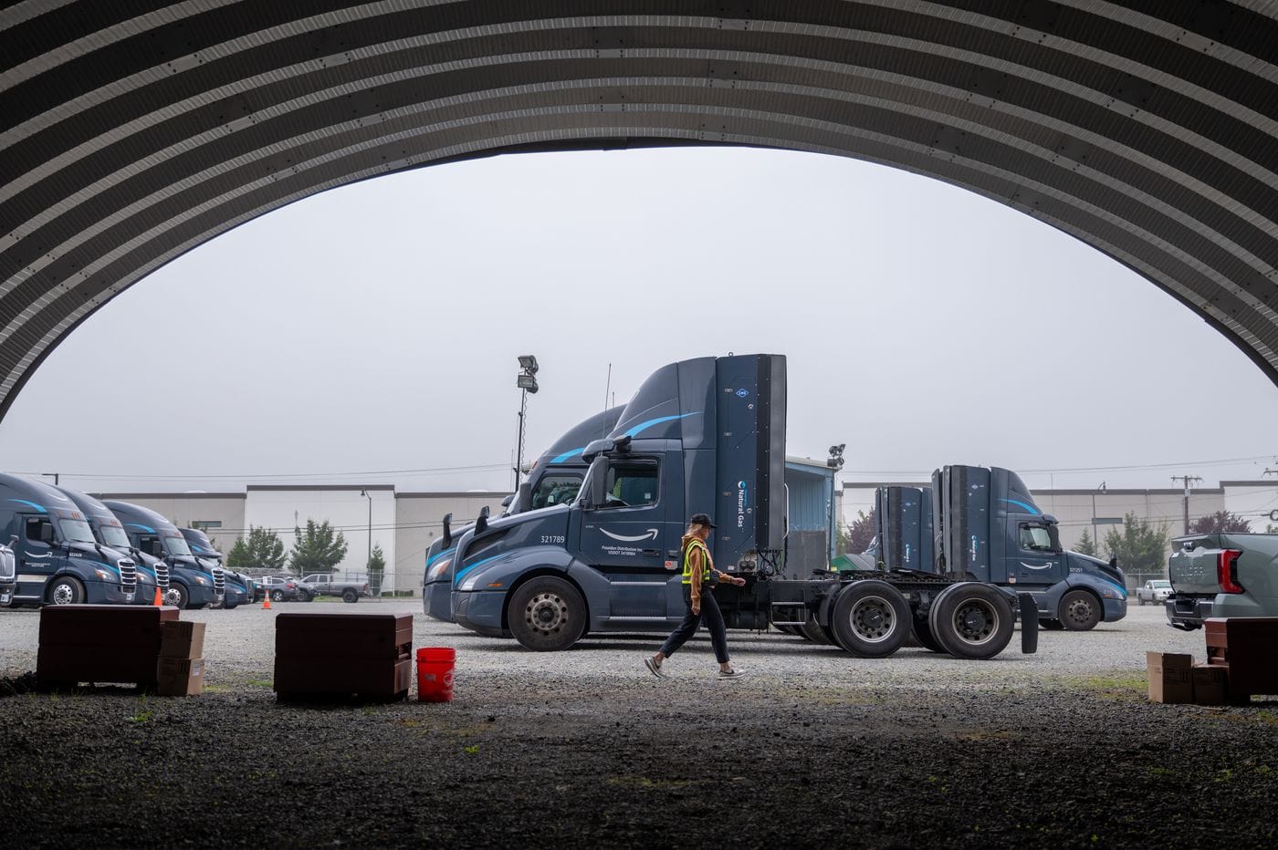 A person in a high-visibility vest walks across a gravel lot in front of several dark grey Amazon semi-trucks under the curved frame of a large metal structure.