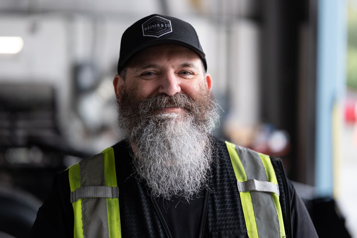 A smiling, bearded man with a graying beard wears a black baseball cap with a Wagner & Co logo and a high-visibility safety vest, standing in an industrial garage setting.
