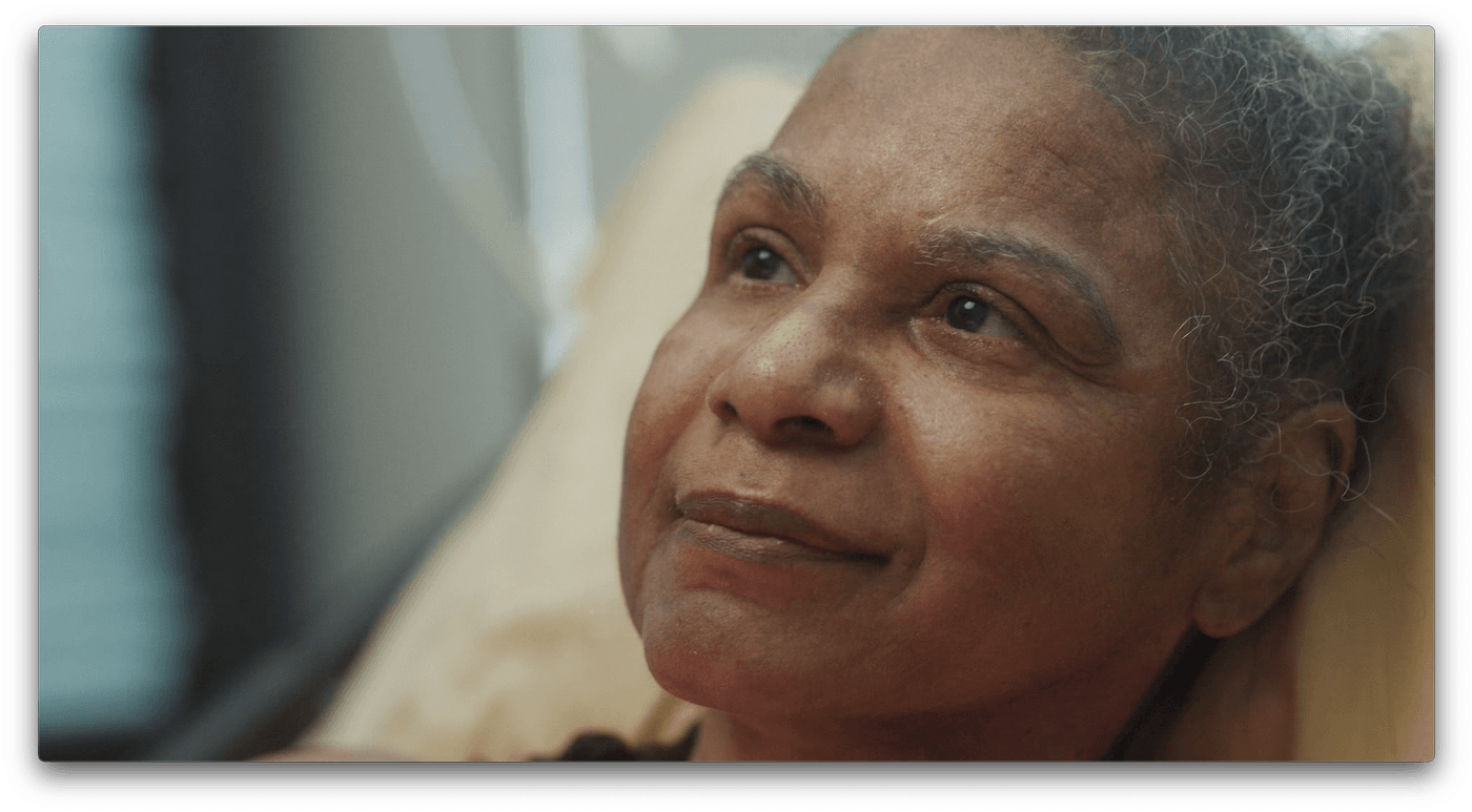 A close-up shot of an older Black woman with short, curly gray hair resting in a hospital bed with a gentle smile while looking upward.