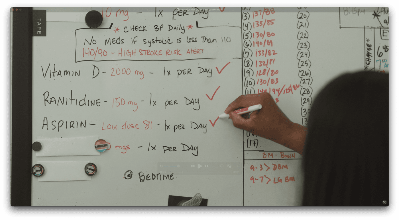 A person uses a red marker to check off a task on a whiteboard listing daily medications, blood pressure tracking data, and bowel movement notes.