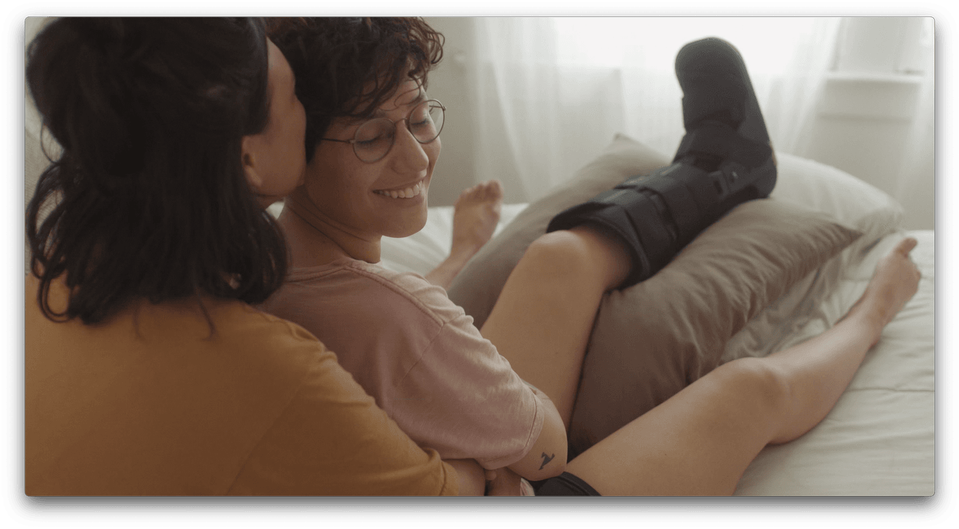 A person wearing a medical walking boot on their lower leg rests on a bed, smiling while a partner sitting behind them kisses their neck.