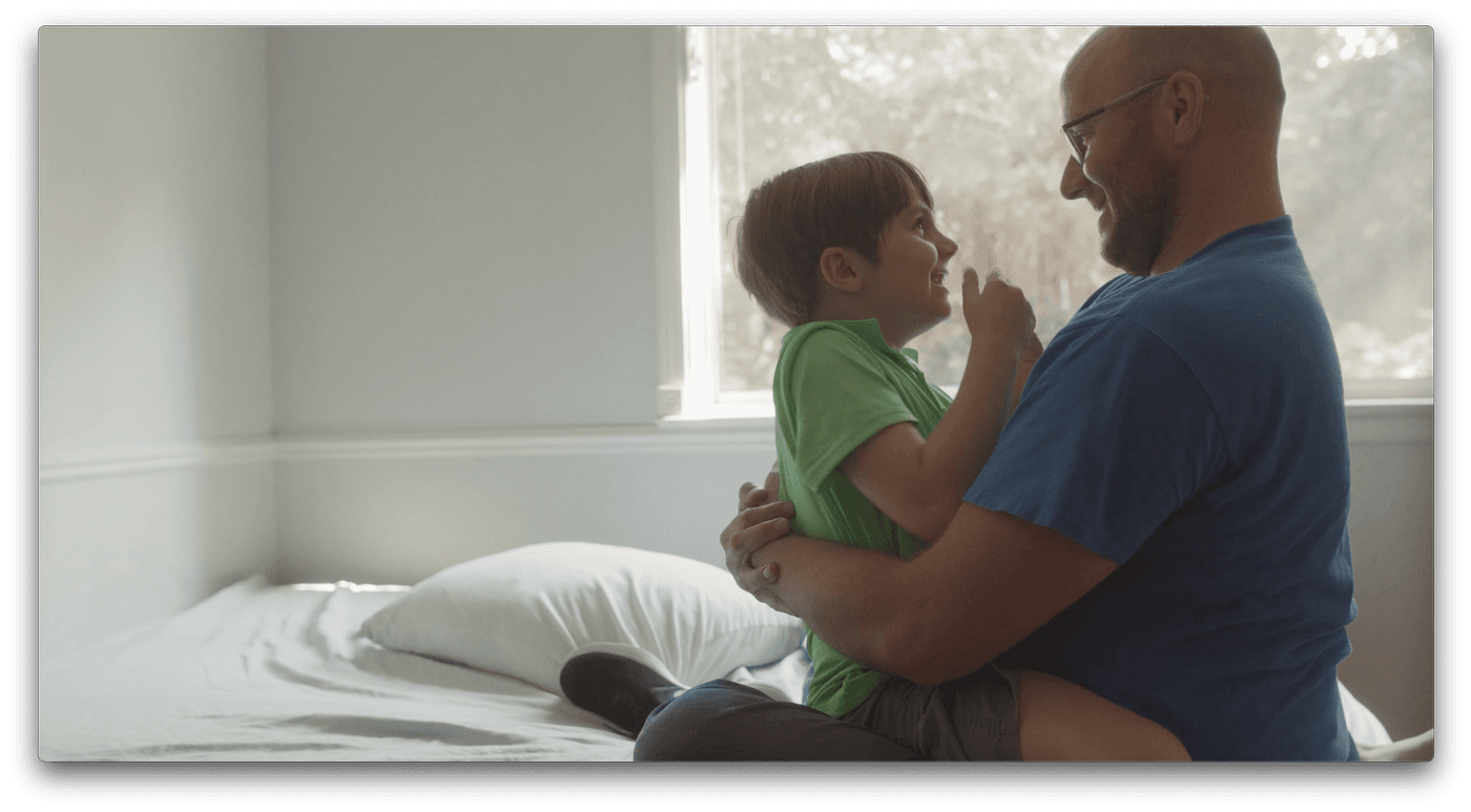 A bald man with glasses and a beard sits on a bed and embraces a young boy wearing a green shirt, both looking at each other with happy expressions.