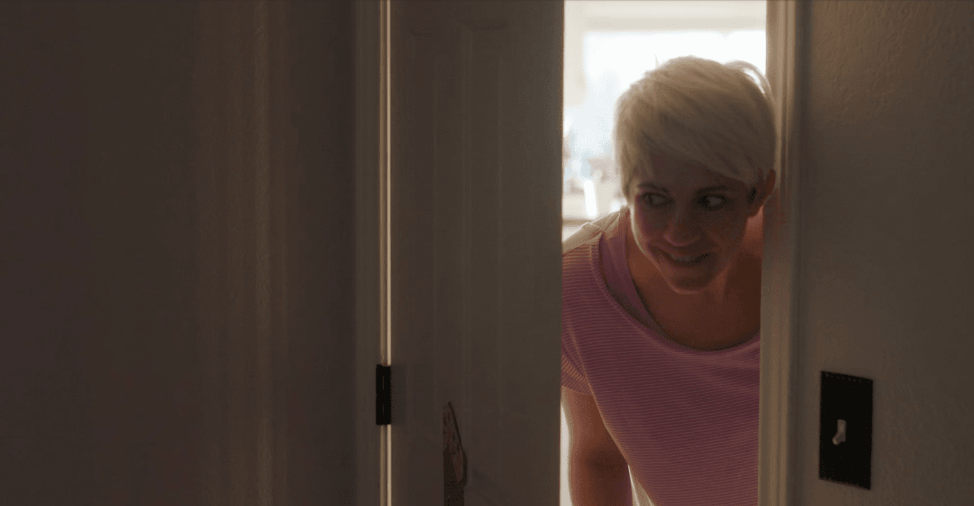 A woman with short blonde hair and a playful expression leans into a doorway, smiling as she peeks into a room.