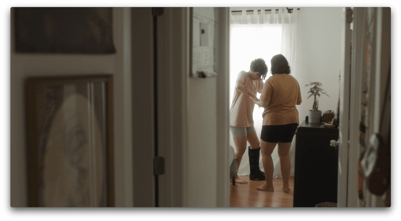 A person in a pink t-shirt helps another person wearing a medical walking boot stand up in a sunlit bedroom.