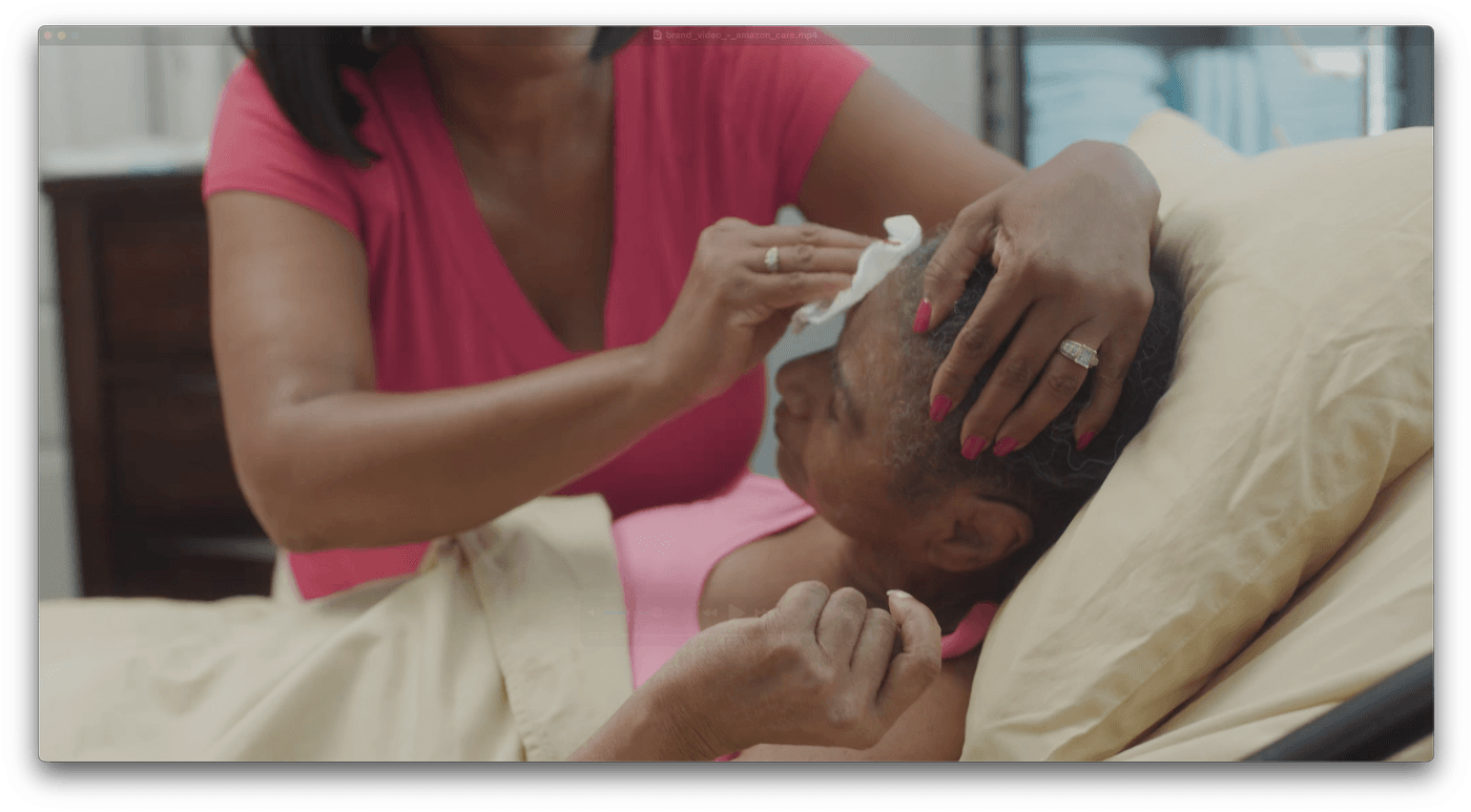 A woman wearing a pink shirt gently wipes the forehead of an elderly woman lying in a hospital bed while holding her head steady.