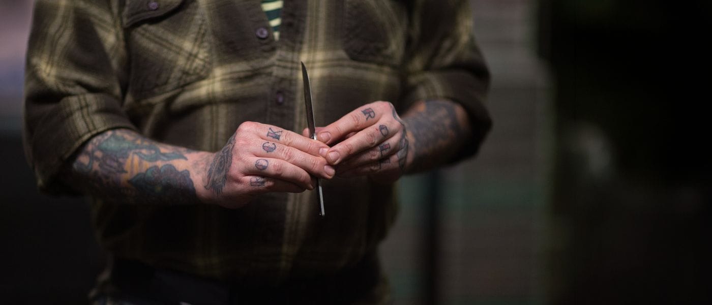 A tattooed person wearing a plaid shirt holds a small folding knife between their hands, which feature knuckle tattoos that read "ROCK" on the left and "ROLL" on the right.
