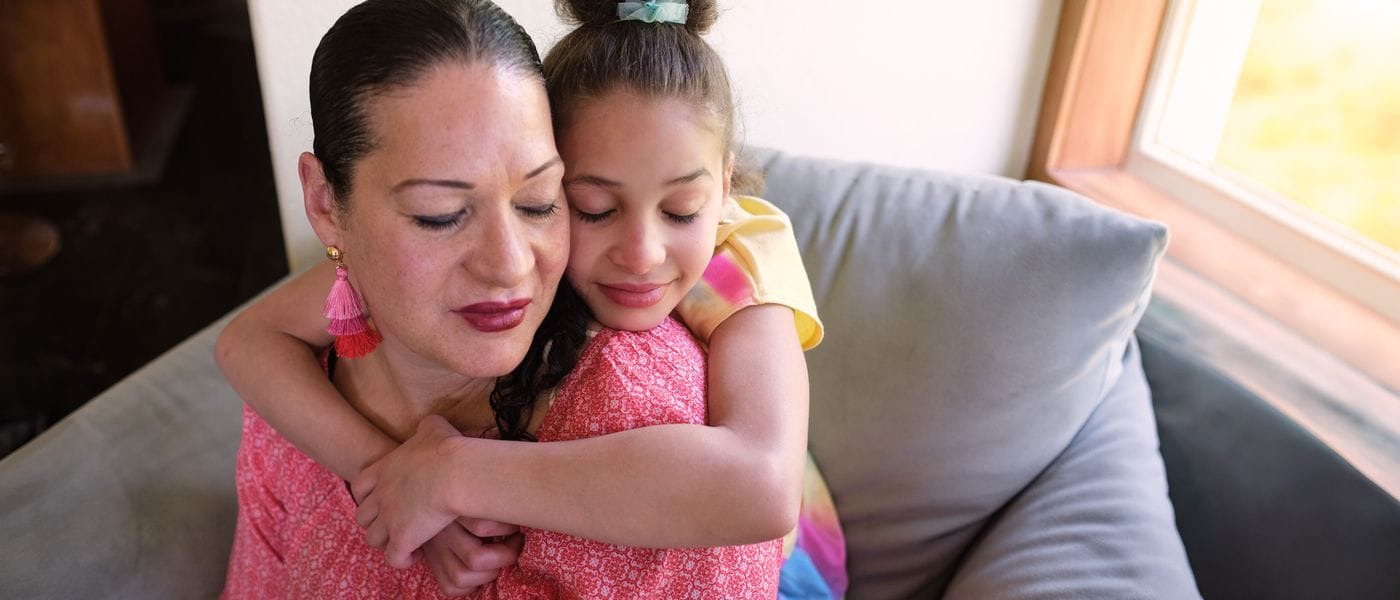 A young girl with her hair in a bun hugs her mother from behind as they both lean their heads against each other with their eyes closed while sitting on a couch.