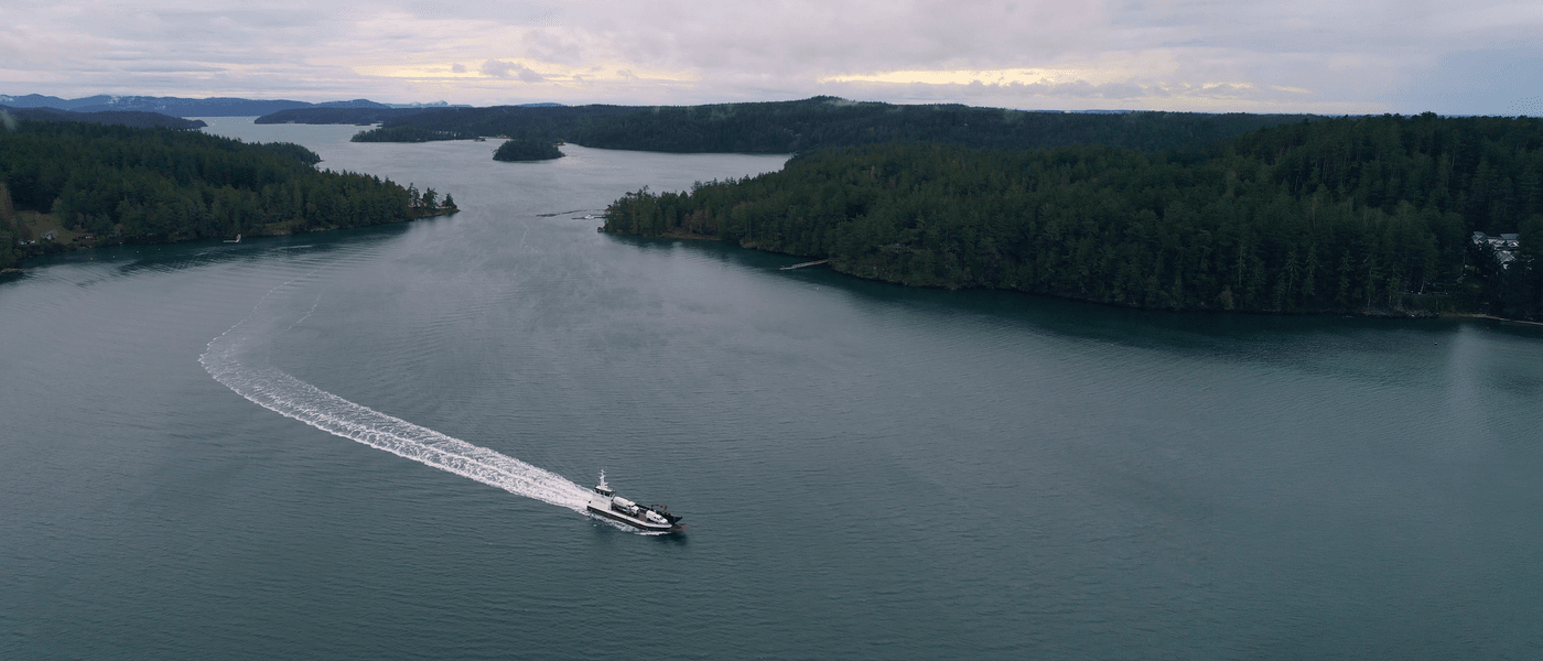 An aerial view shows a small ferry traveling across a narrow, calm channel of deep green water surrounded by dense evergreen forests and rolling hills under a cloudy sky.