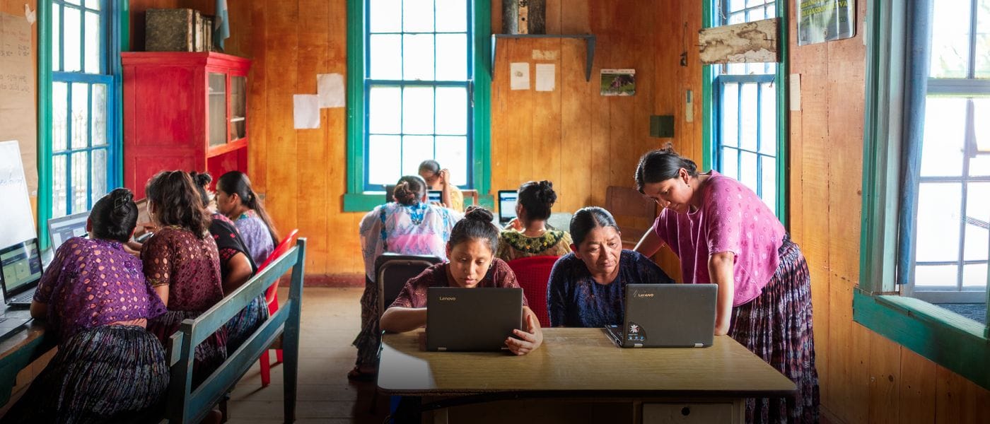 A group of women in traditional clothing work on laptops at wooden tables inside a sunlit classroom, with one woman standing to assist another at a computer.