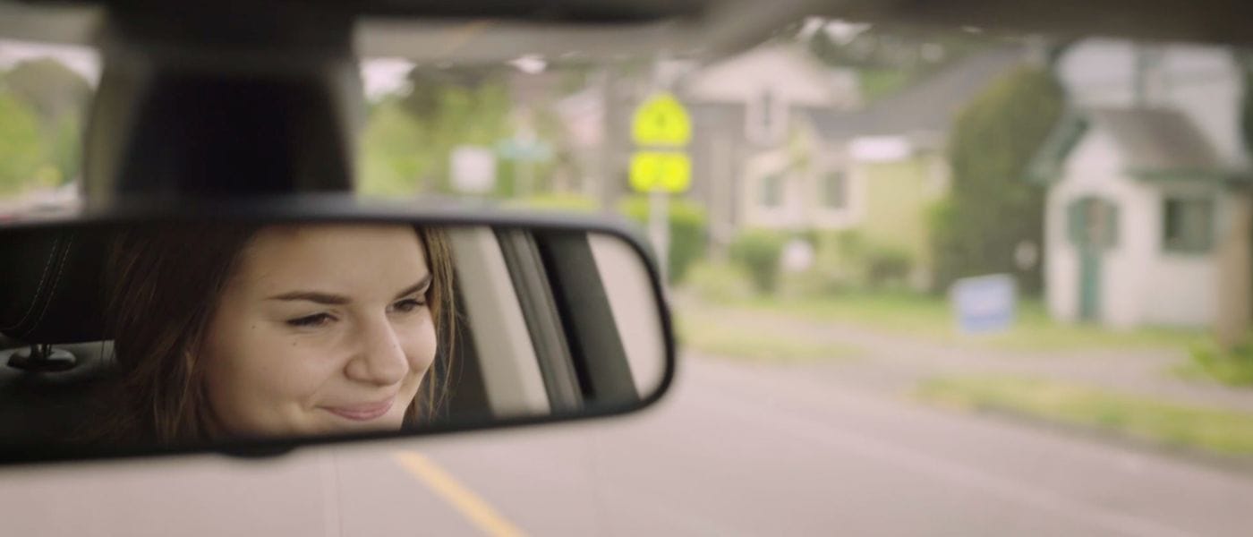 A smiling woman is reflected in the rearview mirror of a car as it travels down a tree-lined residential street.