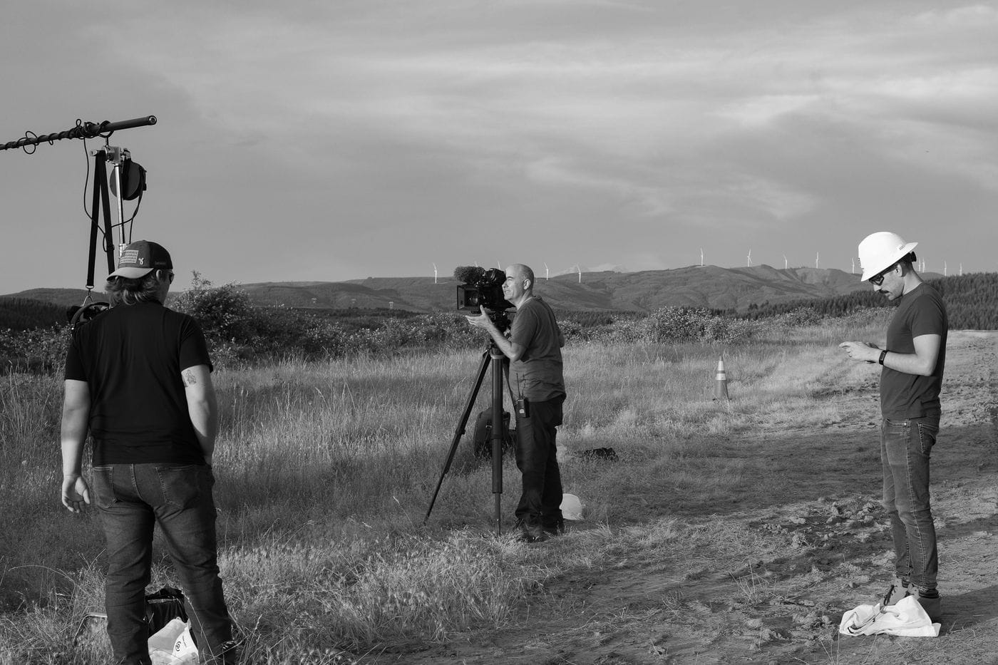 A film crew of three people stands in a grassy field with wind turbines in the background: one man holds a boom mic, another looks through a tripod-mounted camera, and a third wears a white hard hat and looks at his phone.