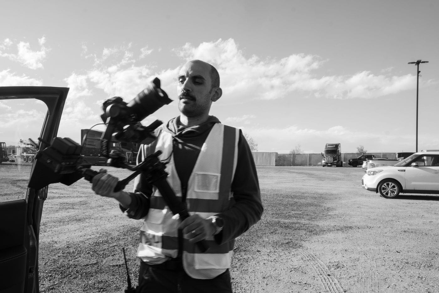 A man with a mustache, wearing a high-visibility vest, stands outside a vehicle in an outdoor lot while holding a camera mounted on a handheld gimbal.