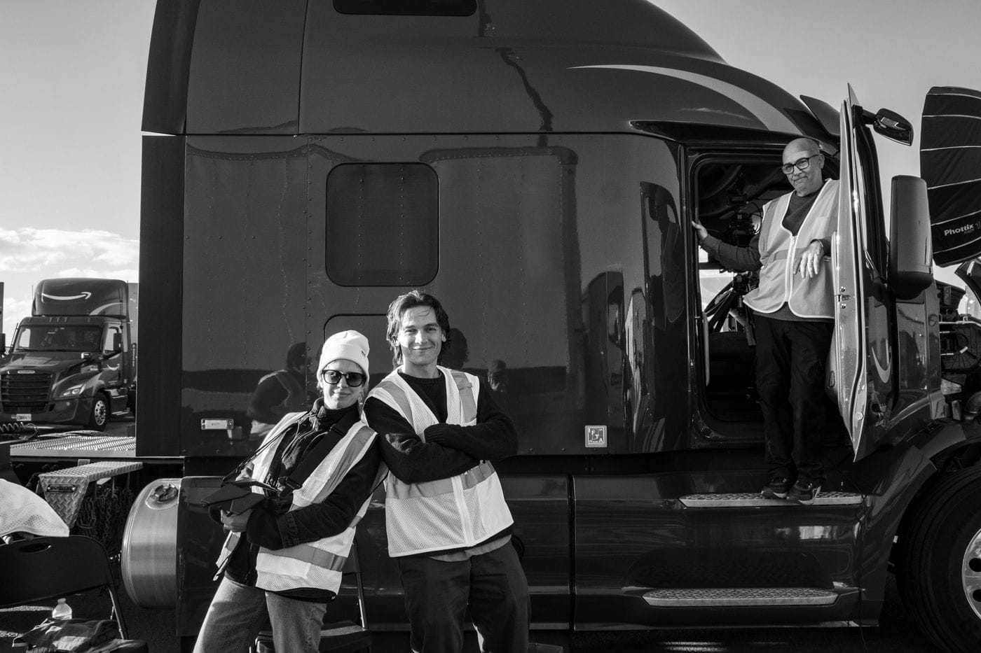 A black-and-white medium shot shows three people standing near a parked semi-truck, with two individuals wearing high-visibility vests posing in front of the vehicle while another person wearing a vest stands in the open cab doorway.