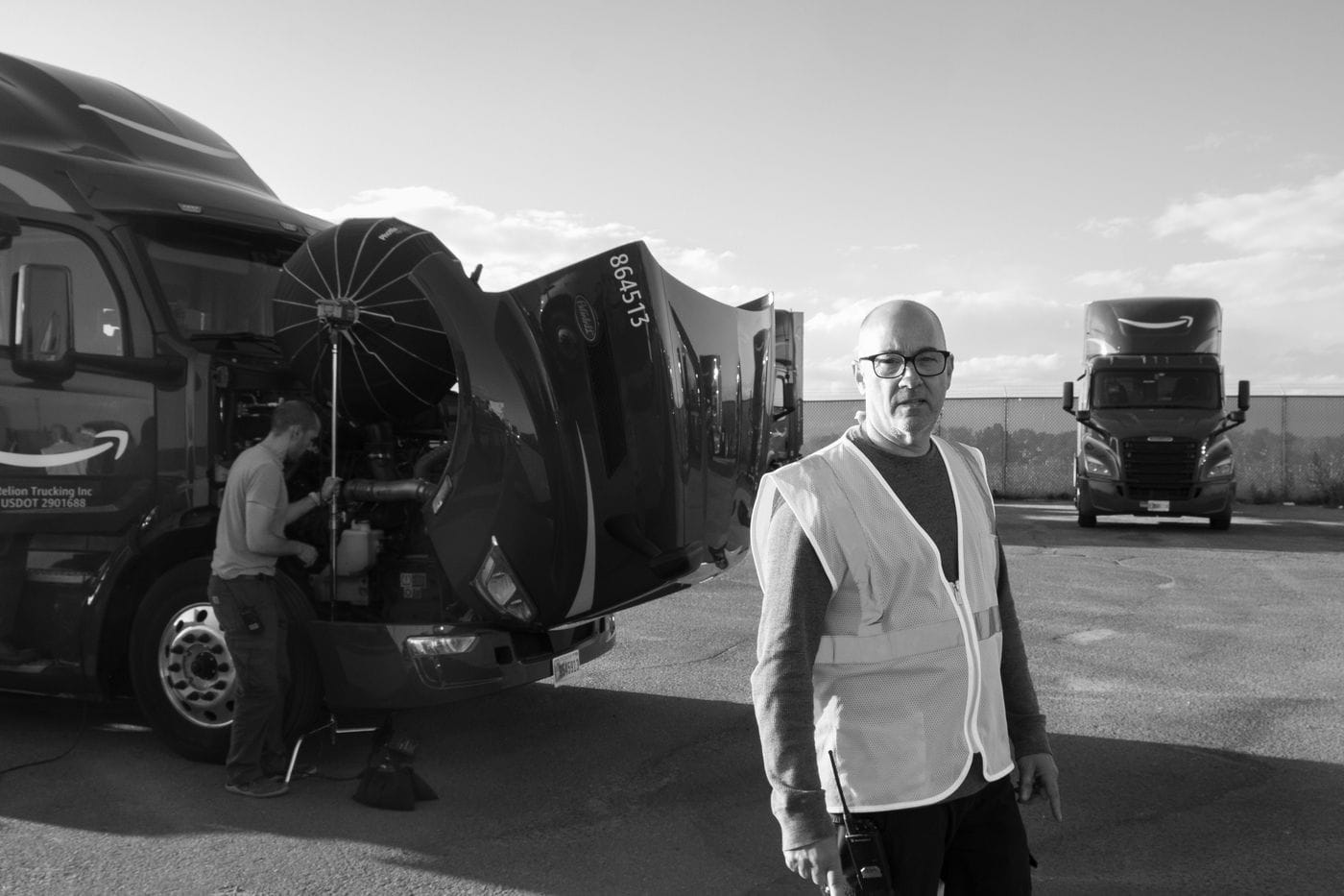 A man in a high-visibility vest stands in the foreground, while behind him a crew member sets up a large lighting modifier over the open engine bay of a semi-truck with an Amazon logo, with another truck parked in the distance.