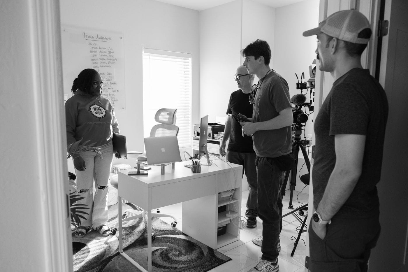 In a brightly lit office, a woman stands by a desk holding a laptop while a film crew with a camera on a tripod prepares to record her.