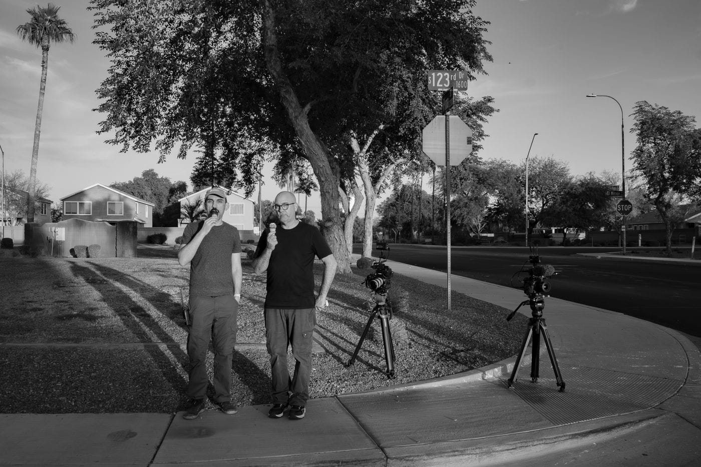 Two men holding ice cream cones stand on a corner sidewalk near a tree and a stop sign, with two professional video cameras on tripods set up nearby.