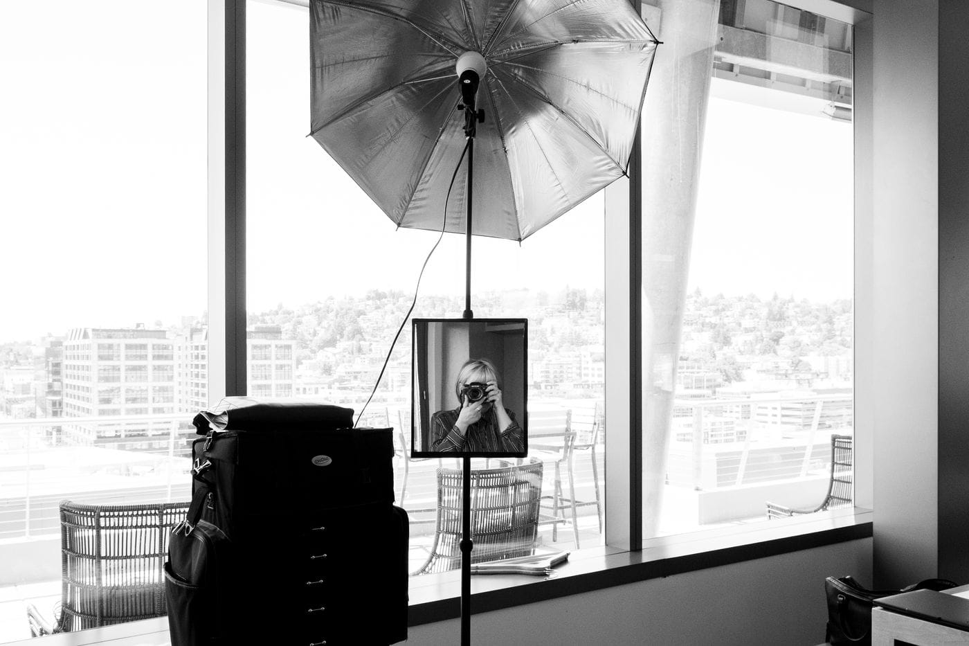 A black and white image shows a professional photography setup with an umbrella light and a mirror attached to a stand, where the photographer is visible holding a camera to take a self-portrait in front of a window overlooking a cityscape.