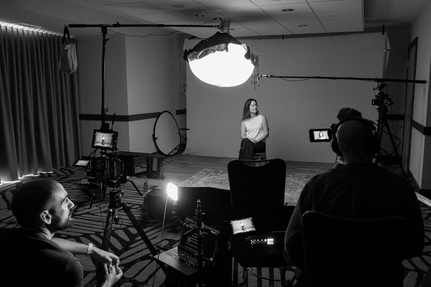 A woman sits on a chair in the center of a room during a film shoot, illuminated by a large softbox light, while a camera operator and director monitor the scene from behind professional production equipment.