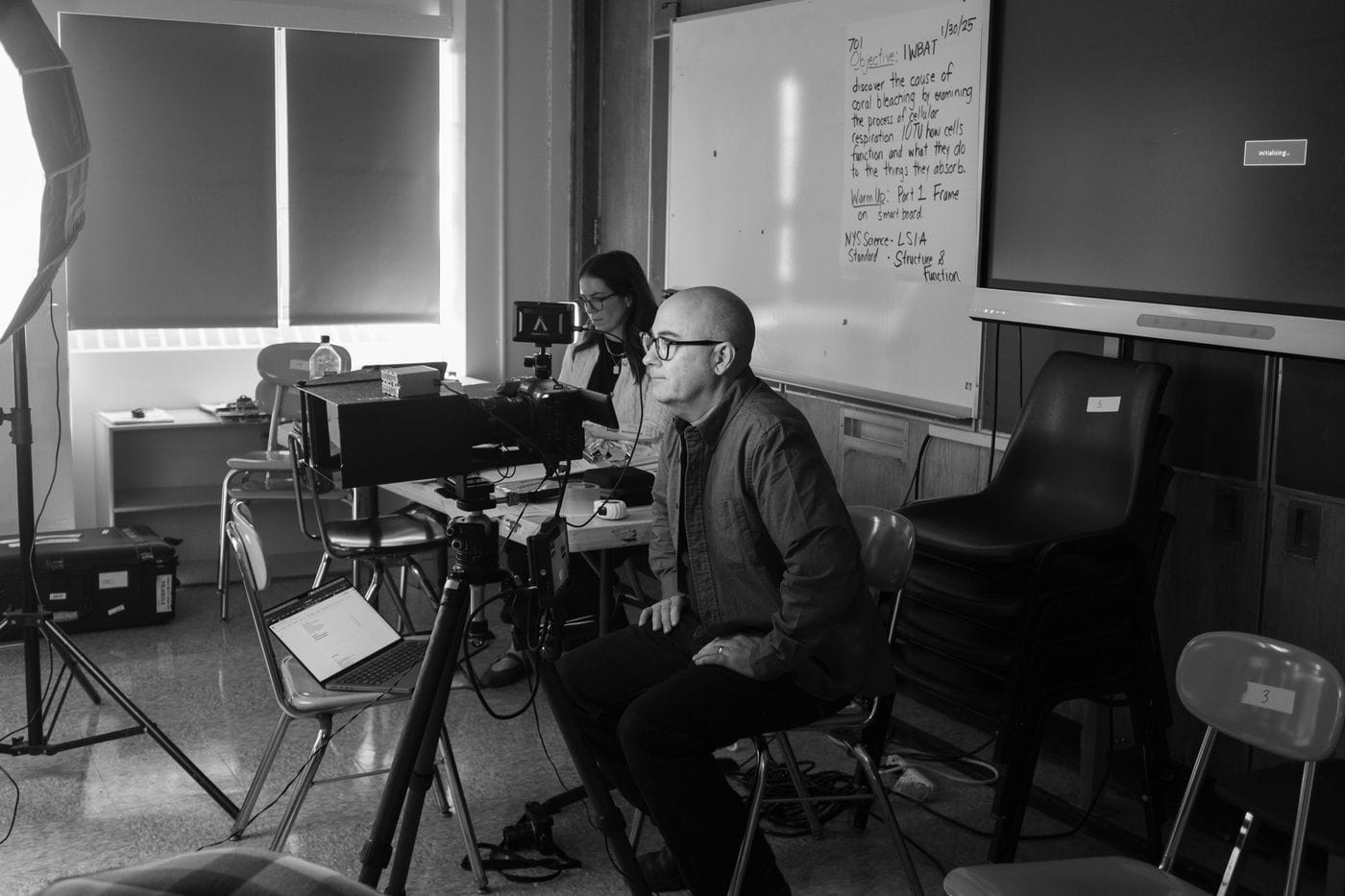 A man with glasses sits in a classroom chair while a camera on a tripod is positioned in front of him, and a woman sits behind a desk in the background in front of a whiteboard with handwritten science lesson objectives.