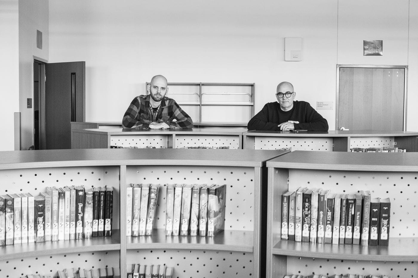 Two men, one younger with a shaved head and plaid shirt and one older with glasses and a black sweater, stand behind library shelves filled with books.