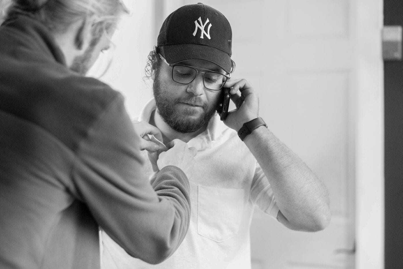 A man in a New York Yankees baseball cap and glasses talks on his phone while another person adjusts his white polo shirt collar.