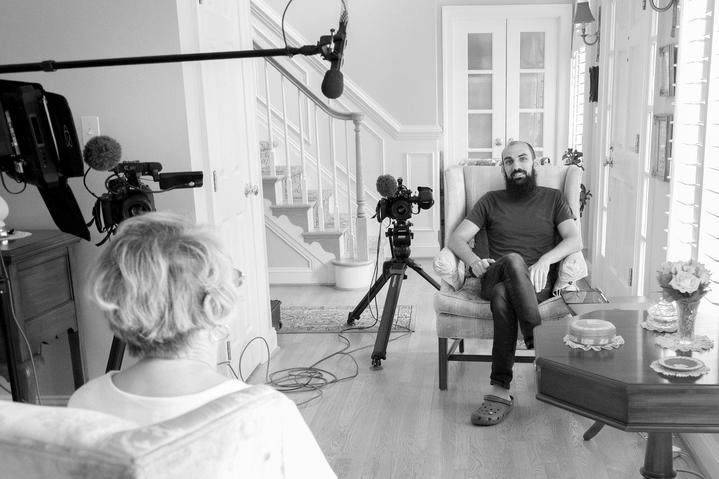 A man with a full beard sits in an armchair while being interviewed by a person seen from the back, with a camera on a tripod and a boom microphone positioned to record the session in a home setting.