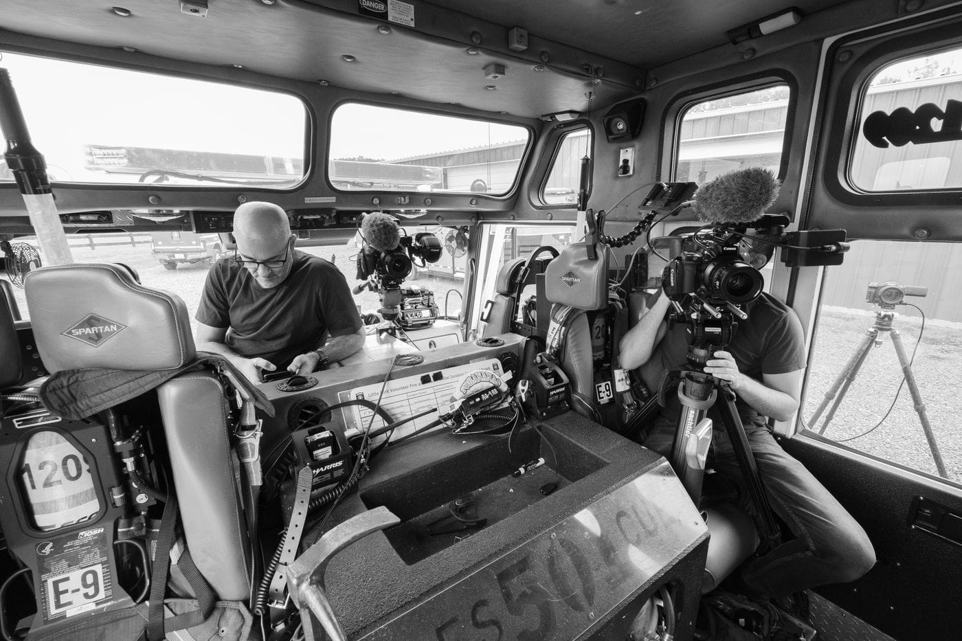 Inside a fire truck cab, a man in a black t-shirt uses his phone while another man operates a professional Canon cinema camera mounted on a tripod.