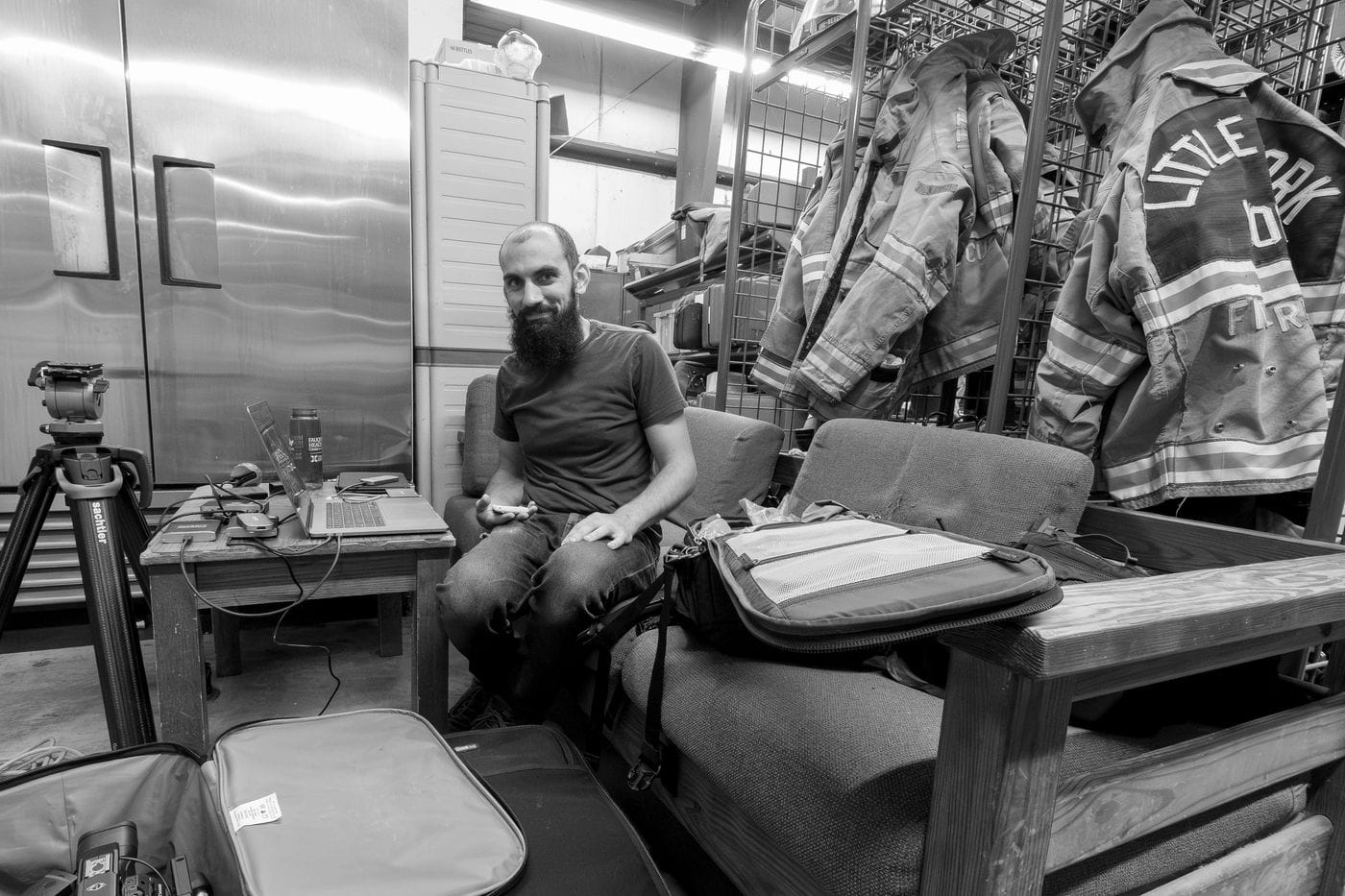 A bearded man in a t-shirt and jeans sits on a sofa in a fire station, looking at the camera while holding a phone, with a laptop on a table nearby and fire gear hanging on the wall behind him.