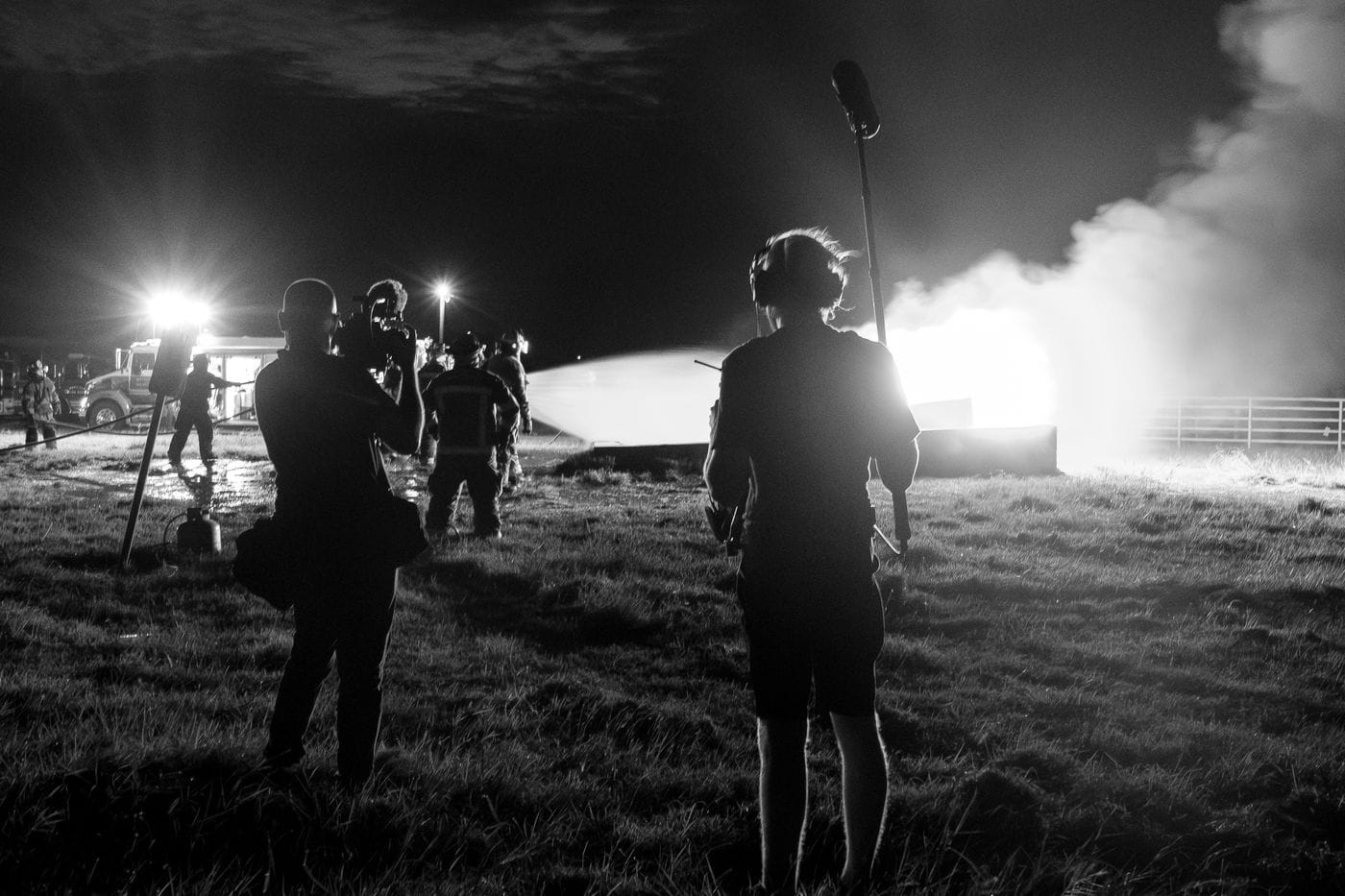 A camera operator and a sound technician stand in a dark field at night, filming firefighters as they spray a bright, intense fire with a hose.