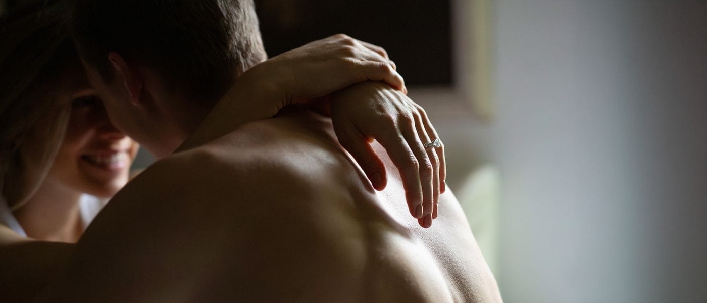 A woman with blonde hair smiles as she embraces a man from behind, her hand resting on his bare back and showcasing a prominent diamond ring.