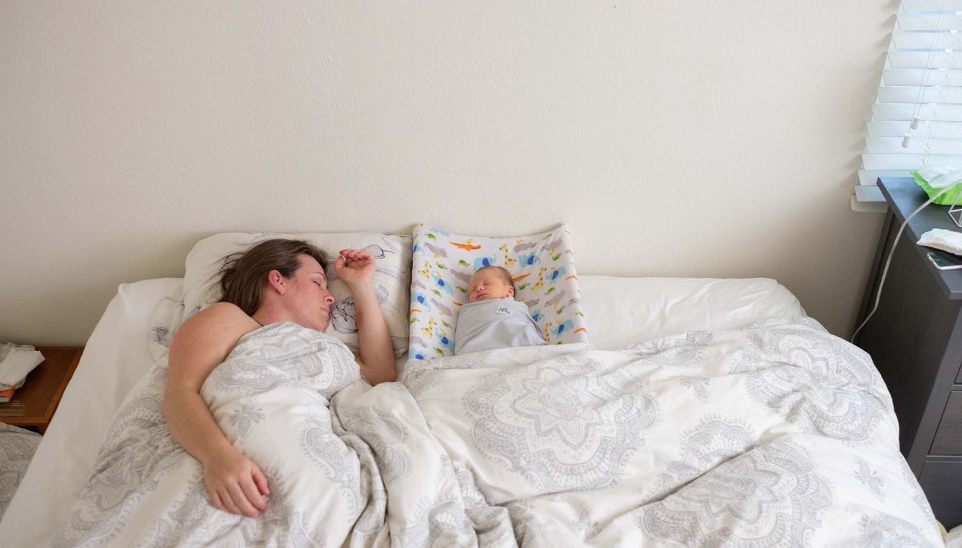 A woman and a swaddled newborn sleep side-by-side in a large bed, with the baby lying on a colorful patterned pad between the pillows.